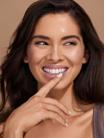 Close-up portrait of a smiling woman with dark hair and glossy mauve lipstick touching her white-manicured fingernail to her teeth.