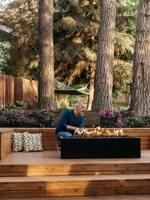 Adult male crouches by a modern black gas fire pit on a wooden deck in a lush backyard setting with tall trees and a hammock.