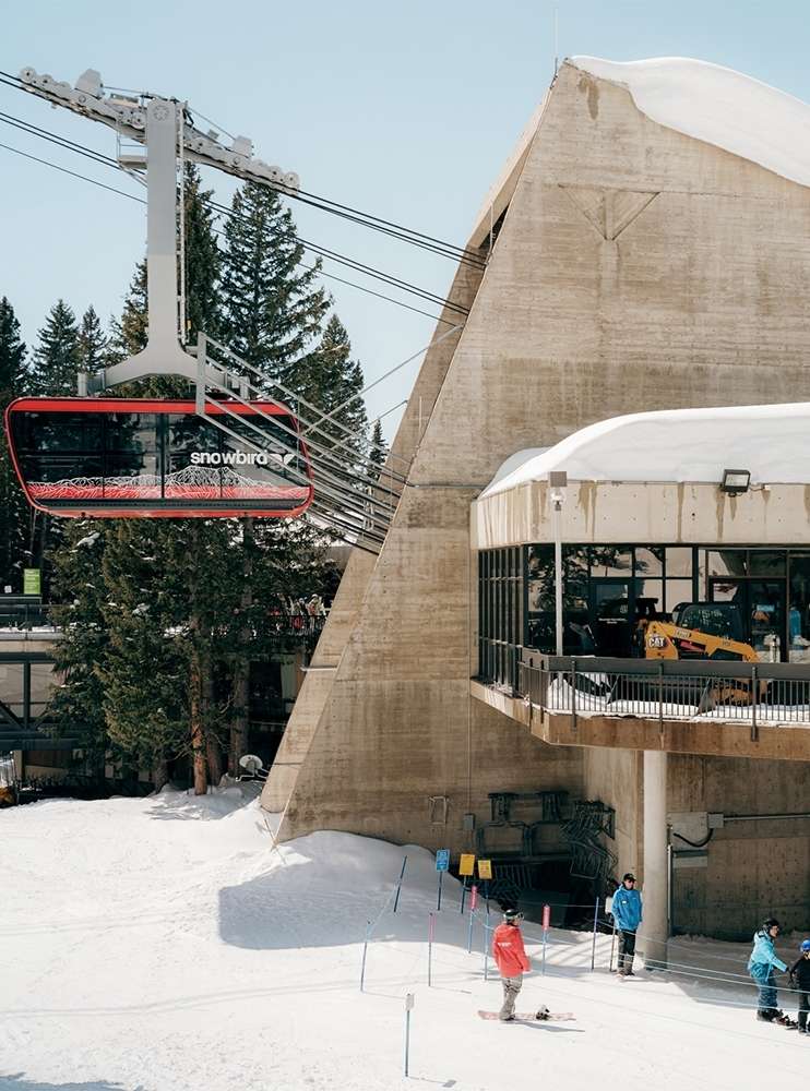 Red Snowbird ski lift ascends mountain at resort with skiers and snowboarders visible in snow covered landscape on a bright day.