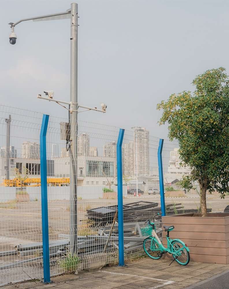 Turquoise bicycle parked next to a fence in an urban setting on a cloudy day, with city buildings in the background.