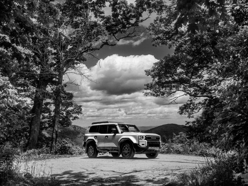 Black and white image of a Toyota Land Cruiser SUV parked on a dirt road surrounded by trees and foliage, with cloudy skies.