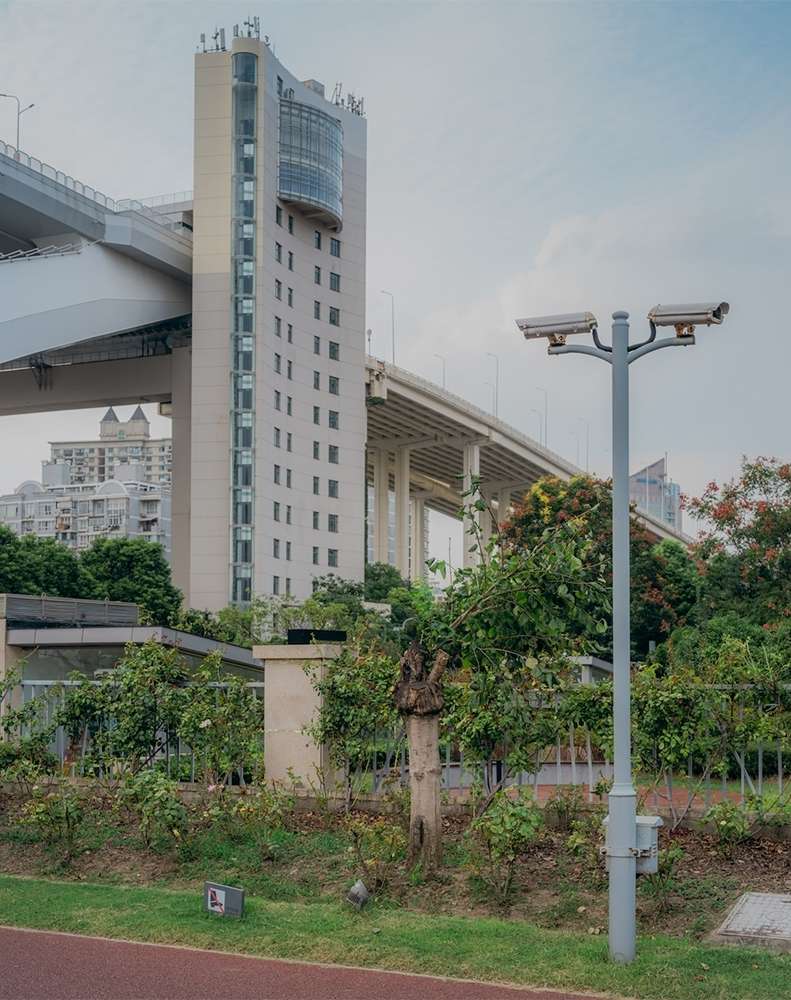 Cityscape featuring a concrete building with windows, an overpass bridge, surveillance cameras, and lush greenery under a cloudy sky.