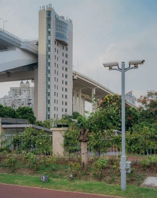 Cityscape featuring a concrete building with windows, an overpass bridge, surveillance cameras, and lush greenery under a cloudy sky.