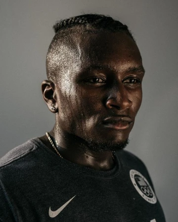 Portrait of a dark-skinned male athlete wearing a dark T-shirt with an Olympics logo and Nike swoosh. He has a shaved head with braided hair.