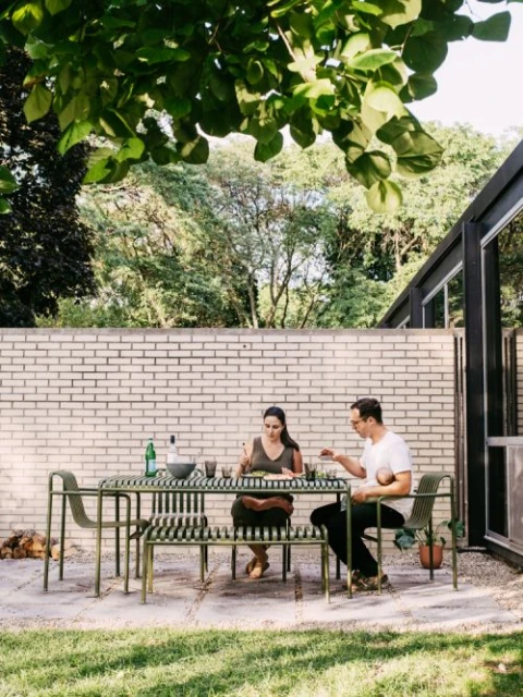 Two adults sit at an outdoor metal table and chairs on a patio near a brick wall and trees, enjoying food in daylight.