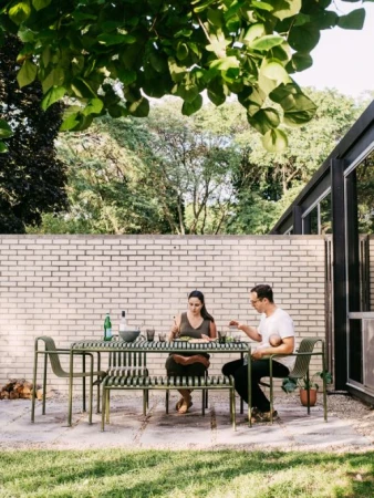 Two adults sit at an outdoor metal table and chairs on a patio near a brick wall and trees, enjoying food in daylight.