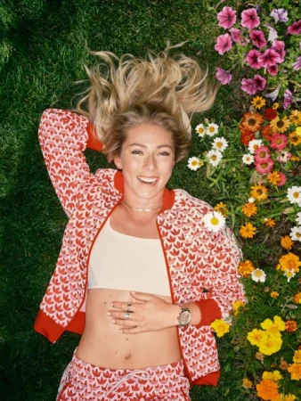 Smiling woman in red and white patterned tracksuit lying on green grass next to colourful flowers, photographed from above.