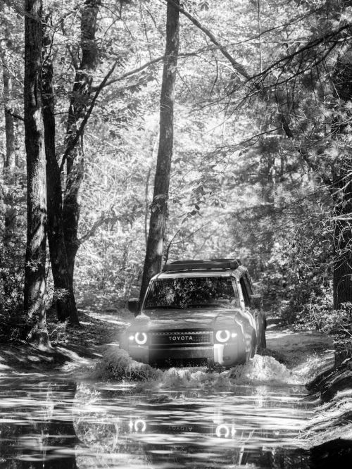 Black and white shot of a Toyota SUV driving through a shallow river in a forest setting with light filtering through the trees.