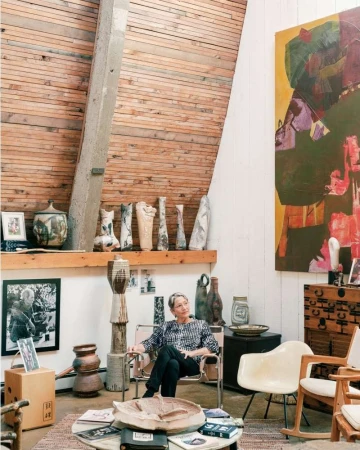 An elderly woman with grey hair seated in a chrome armchair inside an art-filled studio with ceramic sculptures and a large abstract painting.