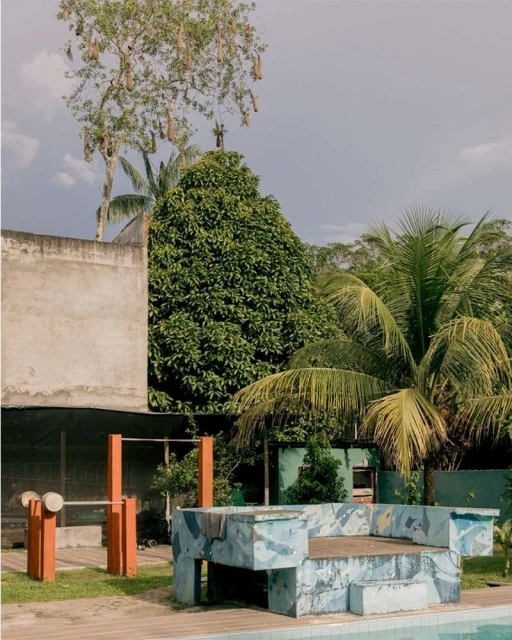 Outdoor scene with a weathered concrete bench near a pool, surrounded by lush greenery, including palms and various tropical trees.