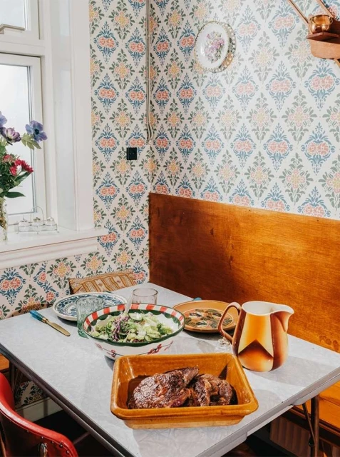 Overhead shot of a dining table set with a bowl of salad, cooked meat, plates, and a pitcher in a cozy, patterned kitchen.