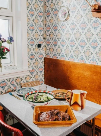 Overhead shot of a dining table set with a bowl of salad, cooked meat, plates, and a pitcher in a cozy, patterned kitchen.