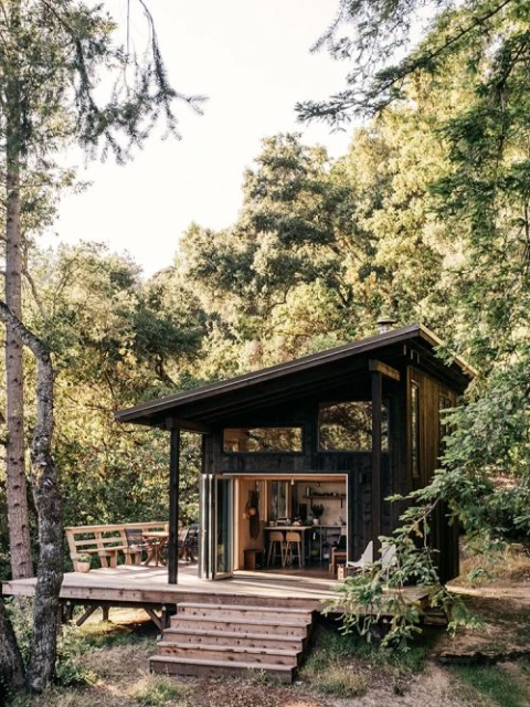 A modern cabin nestled among trees features a wooden deck with outdoor seating and large windows, blending nature and architecture.