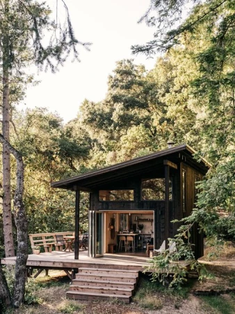 A modern cabin nestled among trees features a wooden deck with outdoor seating and large windows, blending nature and architecture.