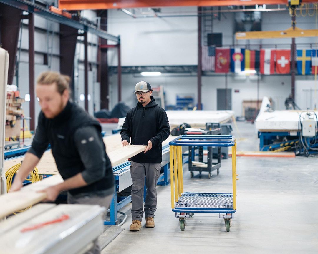Two men working in a factory, one carrying wood, the other at a machine, with flags in the background and a blue and yellow cart.