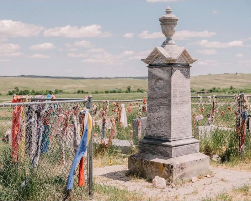Historic marble monument in a grassy field, adorned with colorful ribbons tied to a chain-link fence, set against a cloudy sky.