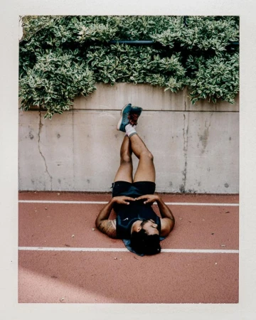 Man lying on a running track with legs resting up against a concrete wall under green foliage