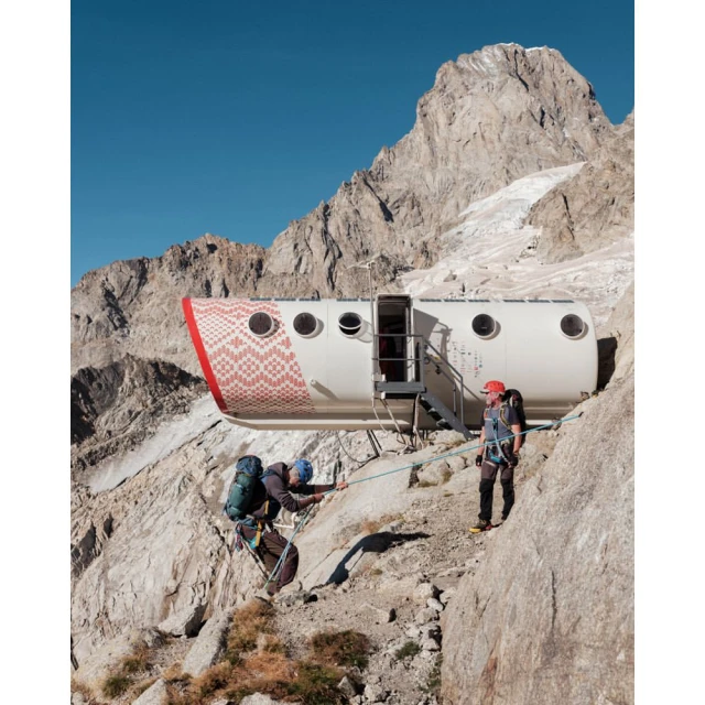 Two mountain climbers with ropes ascending a steep, rocky mountain near a research station, under a clear blue sky on a sunny day.