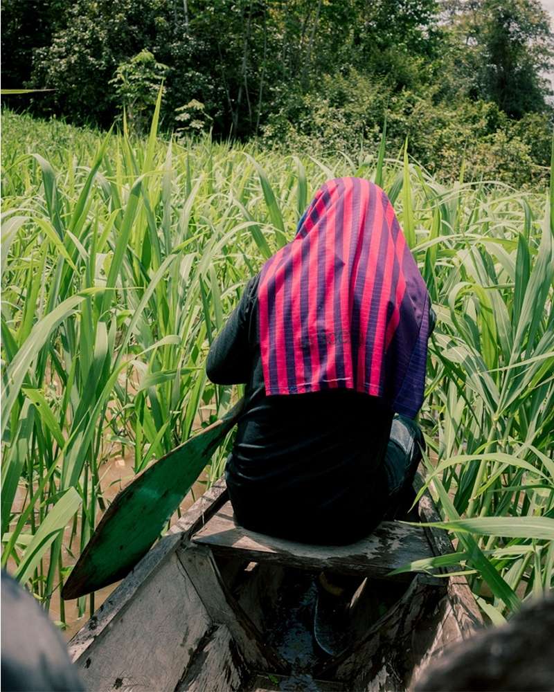 Person in a boat paddles through a waterway surrounded by tall green plants, wearing a striped head covering, in a verdant outdoor setting.