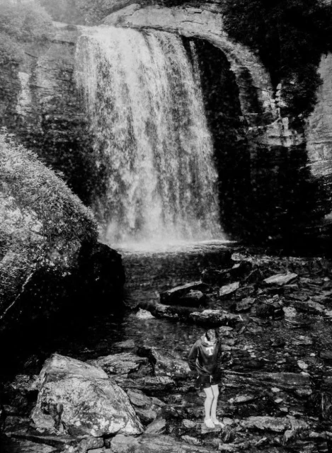 Monochrome image captures a person near a waterfall, framed by rocks and foliage. The scene is high contrast, emphasizing natural textures.
