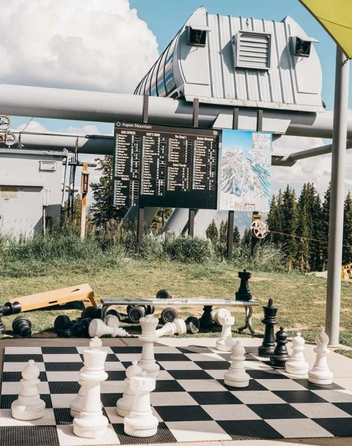 Outdoor chess game with oversized pieces on a black and white board, mountain structure in the background on a sunny day.