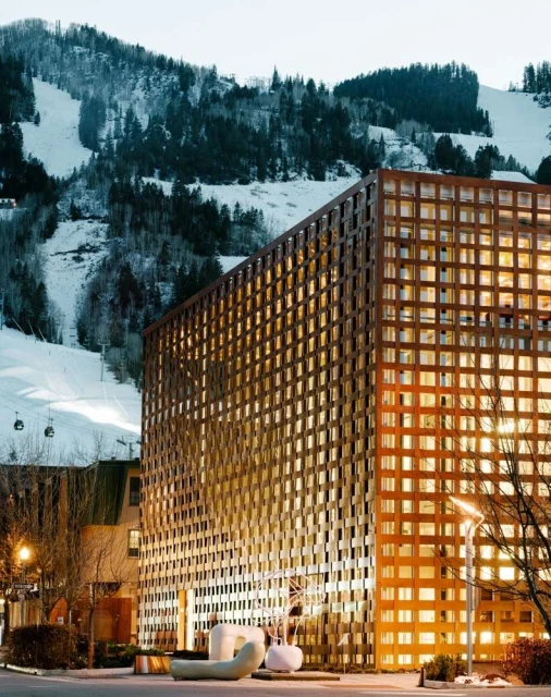 Exterior view of a modern building with a geometric facade against a backdrop of snowy mountains and trees, showcasing architecture in nature.