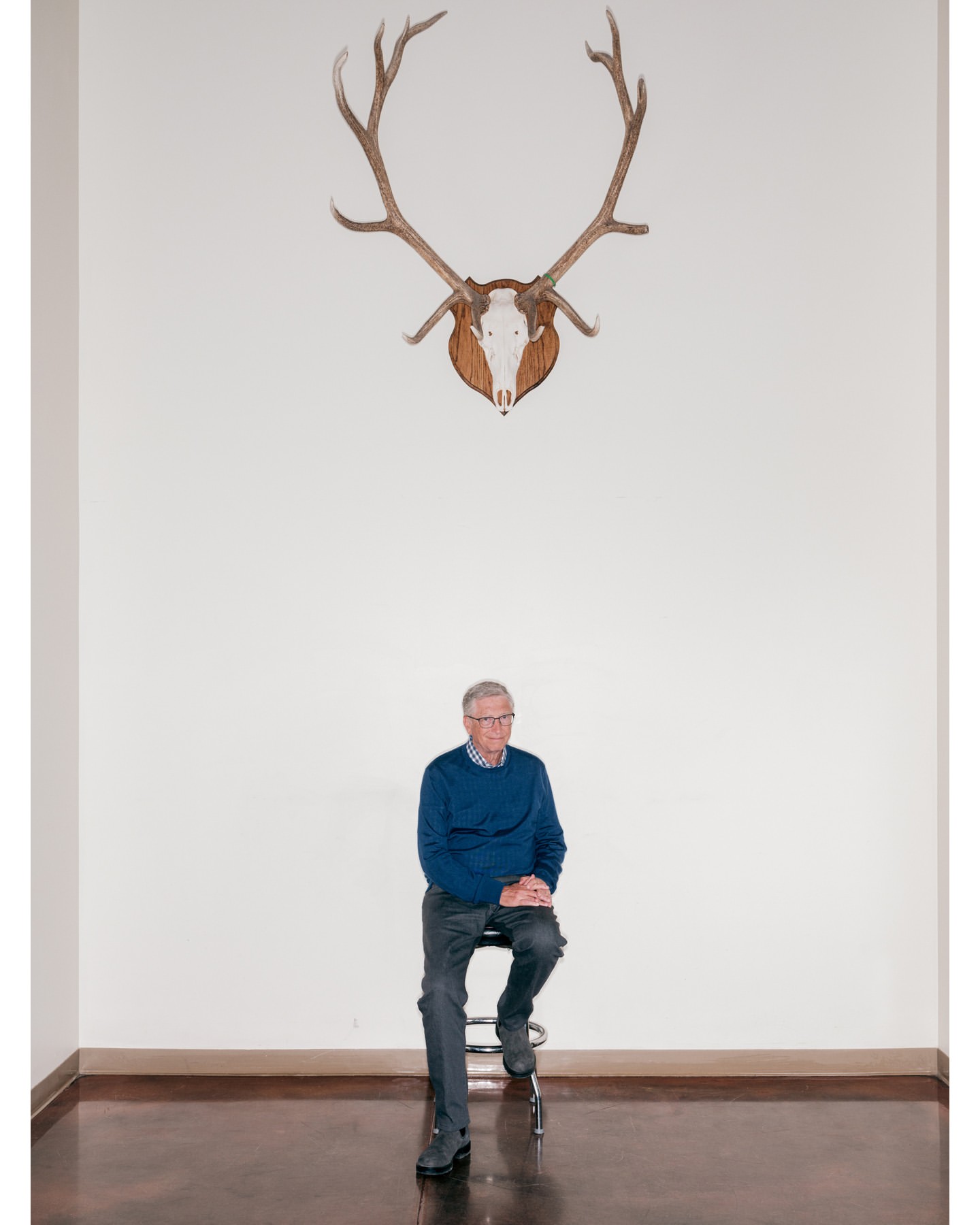 Elderly man sits under mounted deer antlers in a minimalist interior. He wears a blue sweater and gray pants on a wood floor.