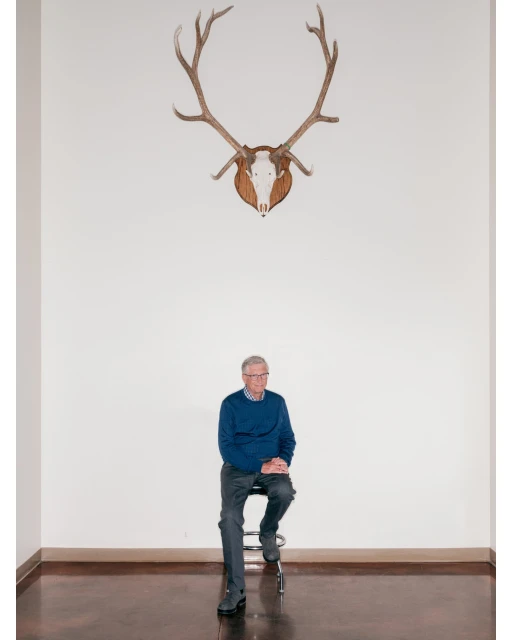 Elderly man sits under mounted deer antlers in a minimalist interior. He wears a blue sweater and gray pants on a wood floor.