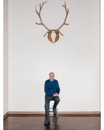 Elderly man sits under mounted deer antlers in a minimalist interior. He wears a blue sweater and gray pants on a wood floor.