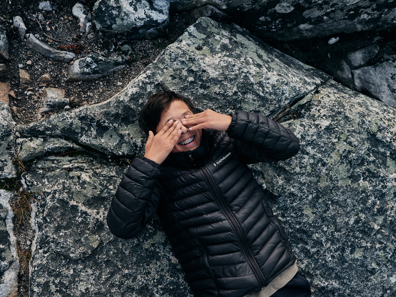 A young person lying on grey rock covering their eyes with their hands, wearing a dark puffer vest.