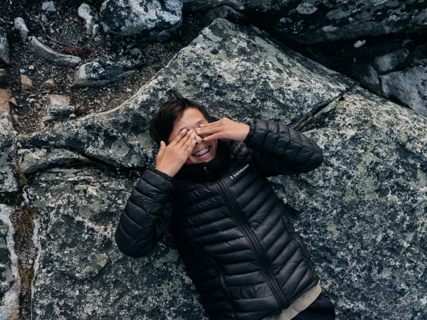 A young person lying on grey rock covering their eyes with their hands, wearing a dark puffer vest.