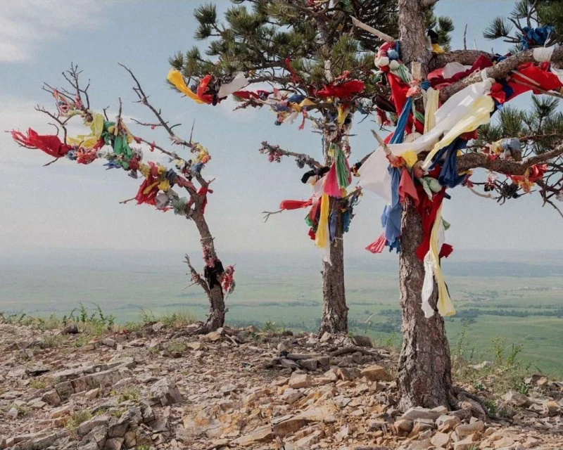 Scenic landscape with three trees adorned with colorful cloths atop a rocky hill overlooking a green valley and pale blue sky.