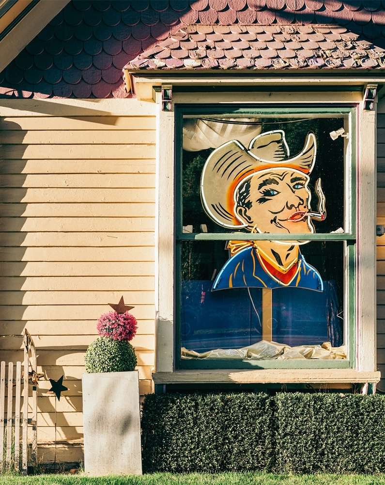 Vintage-style window display features a cutout of a cowboy smoking a cigar, framed by a yellow house facade and green decorative bushes.