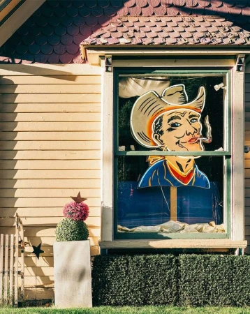 Vintage-style window display features a cutout of a cowboy smoking a cigar, framed by a yellow house facade and green decorative bushes.