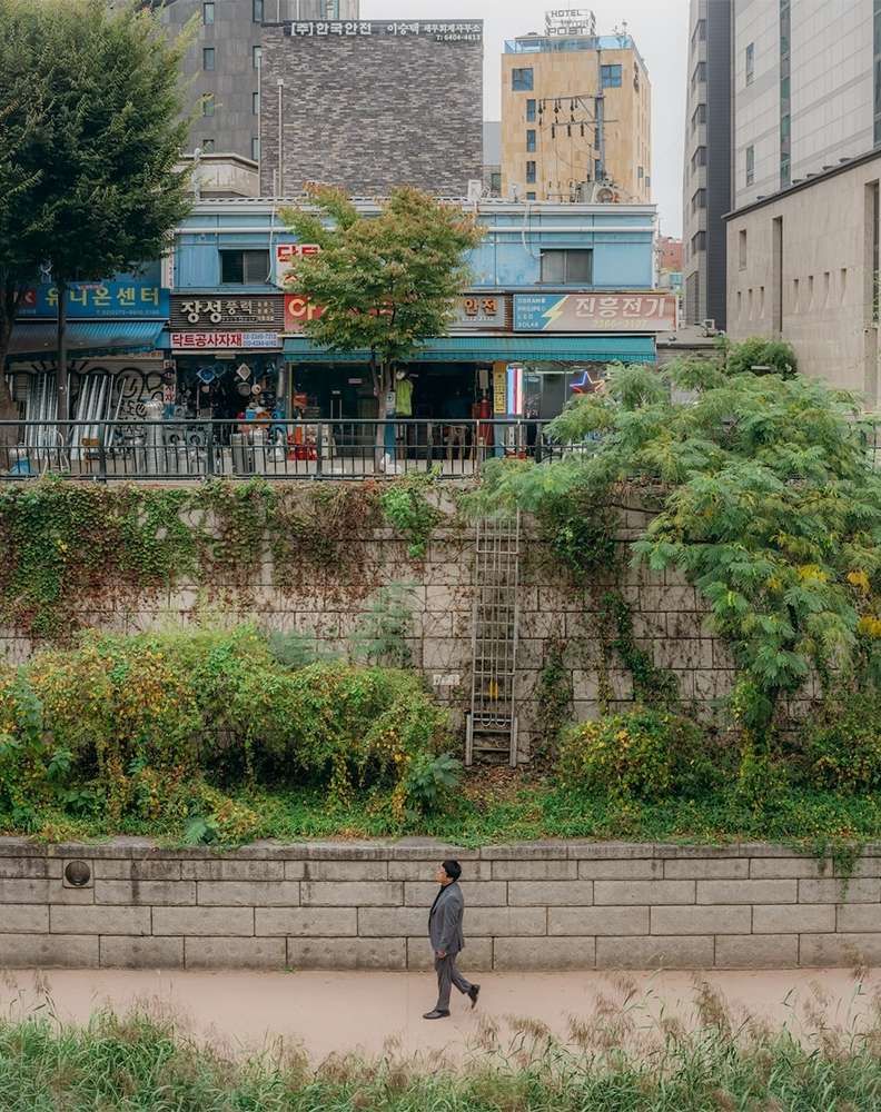 A person in a grey suit walking along a concrete path beside a river lined with lush greenery in an urban cityscape setting.