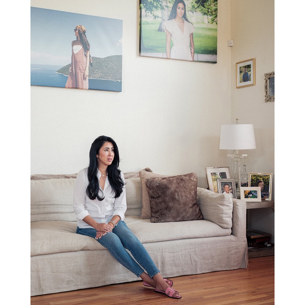 A woman sits on a sofa in a bright living room with artwork hanging on the wall behind her. She's wearing jeans and a white shirt.