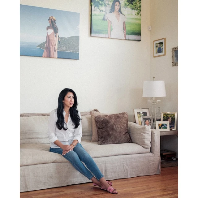 A woman sits on a sofa in a bright living room with artwork hanging on the wall behind her. She's wearing jeans and a white shirt.