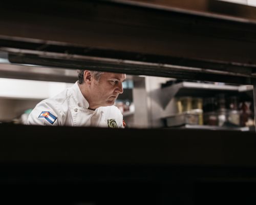 Professional chef in restaurant kitchen wearing white uniform with Colorado flag patch and focused expression, preparing food.
