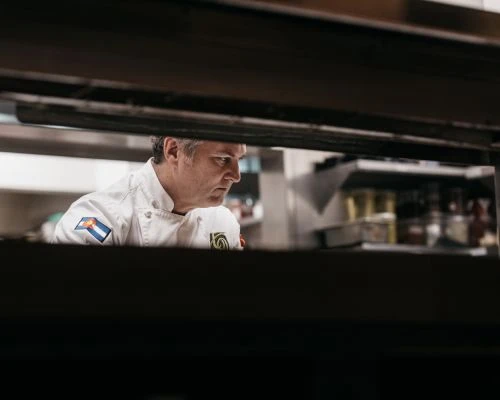 Professional chef in restaurant kitchen wearing white uniform with Colorado flag patch and focused expression, preparing food.