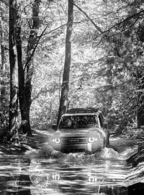 Black and white shot of a Toyota SUV driving through water in a forest, surrounded by trees and reflections of the vehicle.
