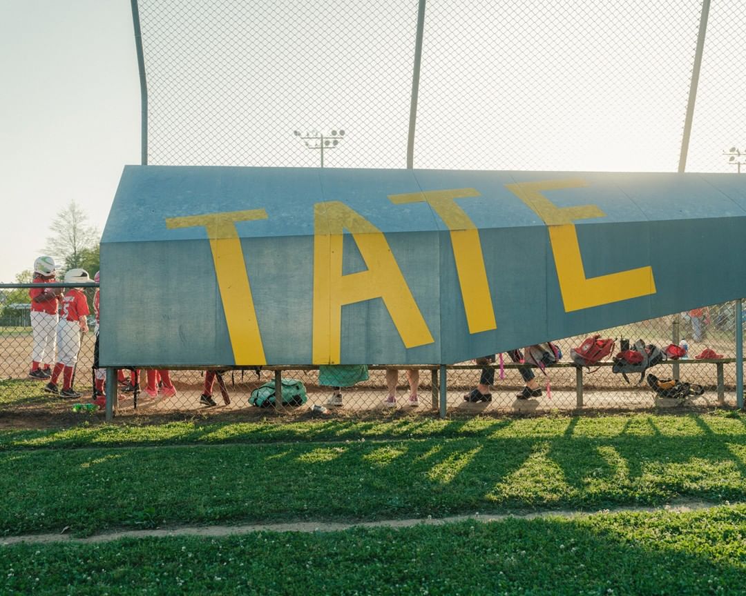 Youth baseball team at dugout with equipment. Chain link fence surrounds a grassy baseball field on a sunny day.