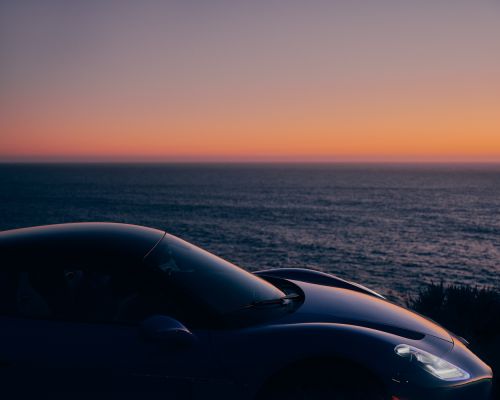 Sleek sports car parked with ocean view at dusk, featuring vivid oranges and pinks from the setting sun in the sky.