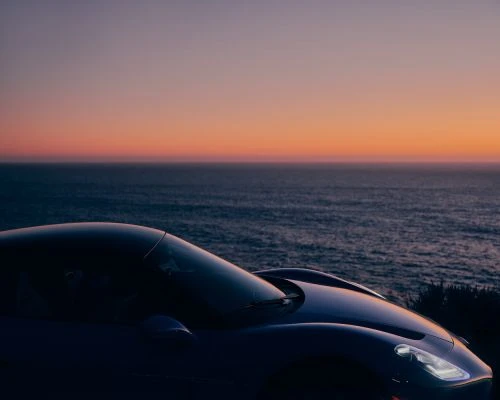 Sleek sports car parked with ocean view at dusk, featuring vivid oranges and pinks from the setting sun in the sky.