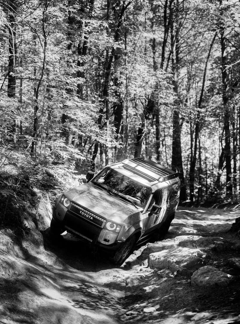 Off-road vehicle, Toyota, drives on a rocky path through a forest. Black and white image with trees surrounding the vehicle.