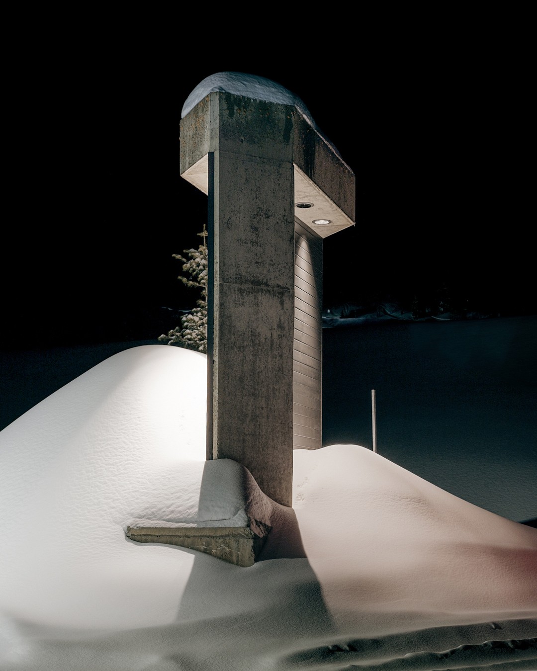 Monochromatic night scene featuring a snow-covered landscape with a modern concrete cross illuminated by hidden lights against a dark sky.