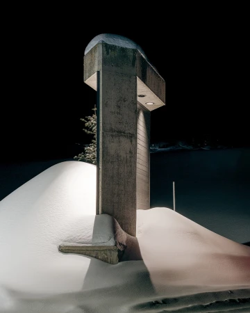 Monochromatic night scene featuring a snow-covered landscape with a modern concrete cross illuminated by hidden lights against a dark sky.