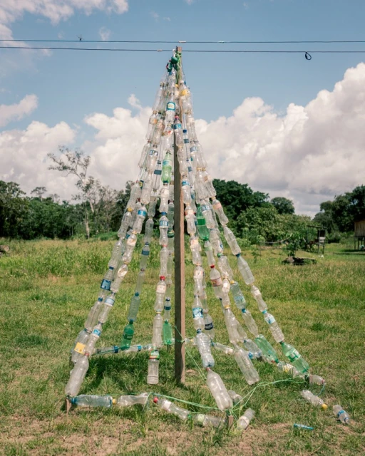 Outdoor shot features a pyramid-like structure crafted from numerous plastic bottles, standing in a grassy field with trees and a blue sky.