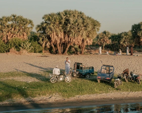 Outdoor scene featuring a group of people, motorcycles, and trucks near palm trees under a bright sky with long shadows.