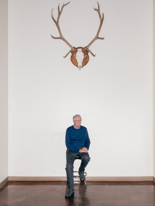 Elderly man in blue sweater seated beneath deer antlers on a stool in a minimalist interior, portrait-style, high-key lighting.