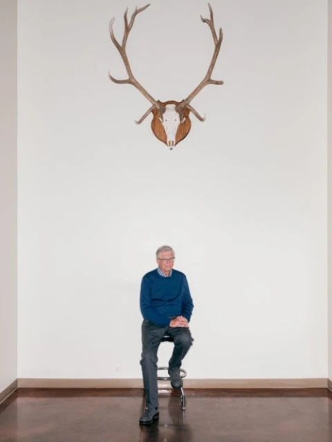 Elderly man in blue sweater seated beneath deer antlers on a stool in a minimalist interior, portrait-style, high-key lighting.